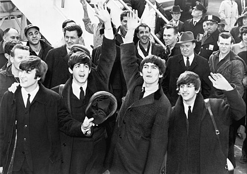 The four members of The Beatles arriving at an airport in 1964, waving to crowds.